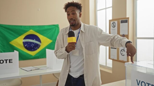 A young man with a beard stands indoors in an electoral college room in brazil, holding a microphone with a brazilian flag in the background.