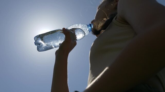 The girl drinks clean, cool water from a bottle. Drink water in the heat against the background of the sun and sky.