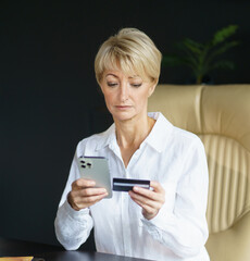 A woman carefully checks her phone while holding a credit card, engaged in an online transaction at her sleek office workspace.