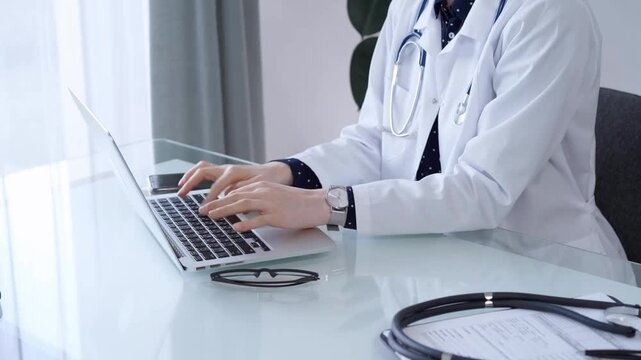 Doctor woman is working with laptop computer at the glass table in clinic. Physician is typing medical information or giving advices online. Medicine and pharmacy