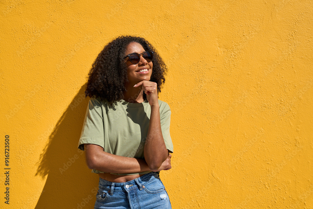 Wall mural Young happy positive African American generation z woman wearing sunglasses smiling looking away at copy space standing at yellow color wall background outdoors feeling joy on sunny summer day.