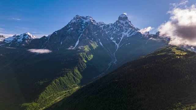 Aerial hyperlapse of Ushba peak in Caucasus mountains during sunrise. Snow-capped mountains, rich green valleys, misty clouds moving fast. Panoramic views for nature, travel, adventure enthusiasts.