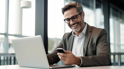 An office worker is smiling while using his phone, sitting at an open desk with a laptop in front of him.