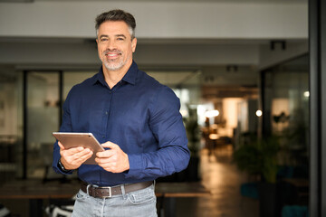 Portrait of happy 50 years old business man executive standing in office using digital tablet. Middle aged businessman manager business owner working on professional finances project looking at camera