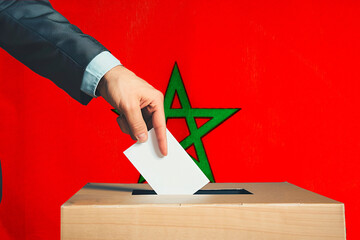 A hand holds a ballot in a ballot box against the background of the Moroccan flag. People place...