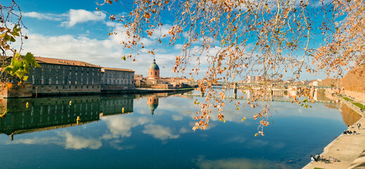 La Grave Hospital Reflected in the Garonne River at Sunrise, Framed by Lush Foliage in the Foreground and Bathed in Golden Light Under a Clear Blue Sky, Toulouse