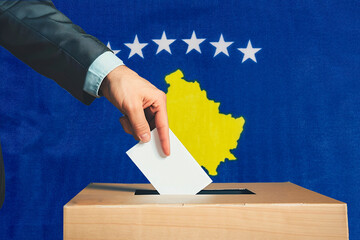 A hand holds a ballot in a ballot box against the background of the Kosovo flag. People place...