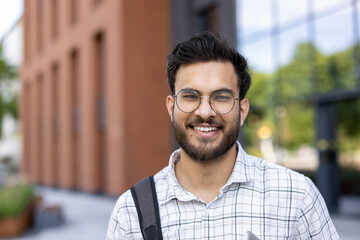 Smiling young man with glasses and beard holding phone outside modern building