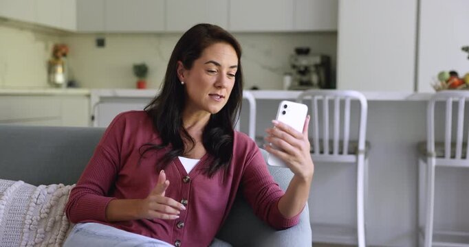 Attractive young Hispanic woman sitting on comfortable couch lead pleasant personal talk with family using smartphone and video call app, share news, enjoy remote conversation on-line. Virtual meeting