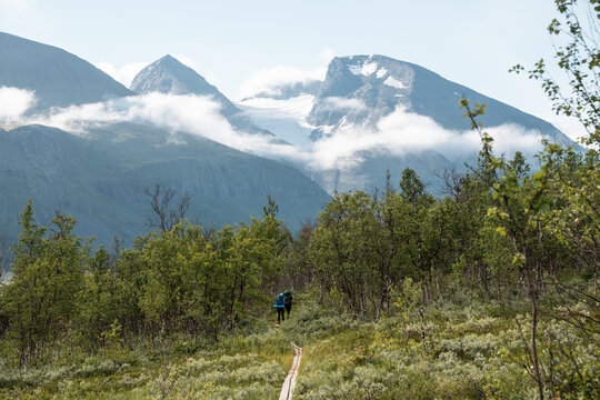 2 Hikers from behind walking a path (Padjelantaleden) in Swedish Lapland National Park. Blue sky and Mount Akka in mystical clouds. Moore and birch tree forest in front.