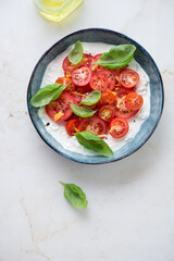 Bowl of stracciatella served with sliced tomatoes and basil, top view on a light-beige marble background, vertical shot with space