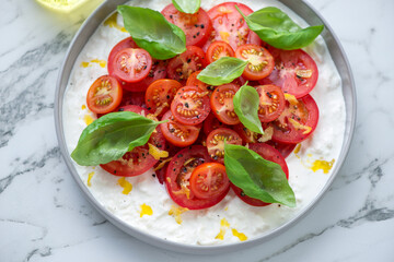 Plate with stracciatella or stretched curd fresh cheese, red tomatoes and green basil, horizontal shot on a white marble surface, middle close-up