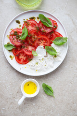 Red tomatoes served with basil oil, burrata and fresh basil, above view on a beige stone background, vertical shot