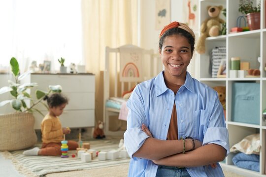 Biracial woman smiling at camera with arms crossed in modern nursery setting Child playing with toys in background and shelves filled with children's items