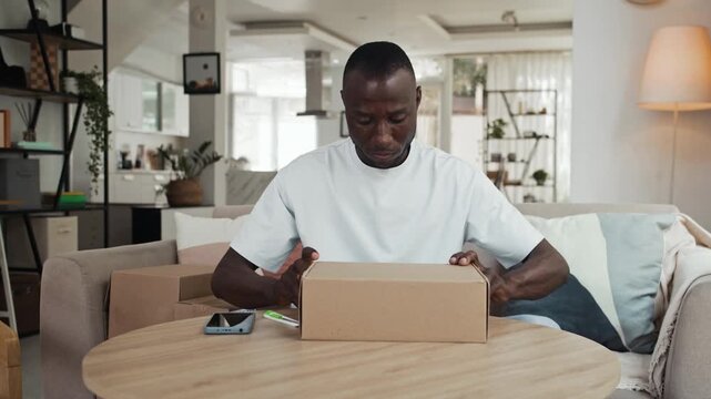 Medium shot of happy black male buyer getting parcel with leather shoes while sitting at table at home