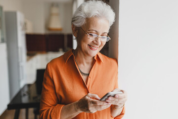 Portrait of cheerful grandma holding smartphone in hands, chatting with friends, browsing social media, watching memes, funny videos standing against wall next to window at kitchen