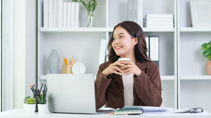 Young businesswoman in a brown blazer and beige sweater sits at her home office desk, smiling and enjoying a cup of coffee.