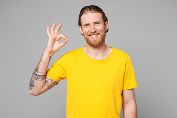 Young smiling happy cheerful fun cool Caucasian man he wearing yellow t-shirt casual clothes look camera showing okay ok gesture isolated on plain grey background studio portrait. Lifestyle concept.