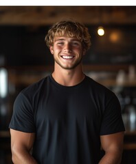 Fototapeta premium Smiling young Caucasian man with short blonde hair wearing a black t-shirt, standing confidently in a warmly lit environment.