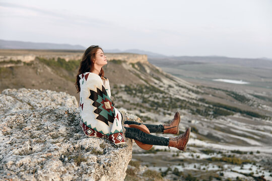 Woman in blanket and boots sitting on rock gazes at stunning mountain and valley vista