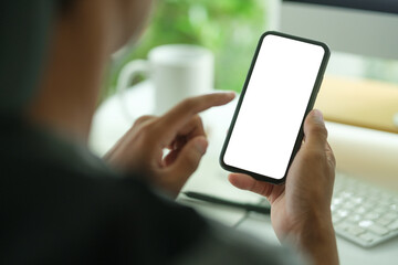 Man using smart phone while sitting front of laptop at office desk. Over shoulder view.