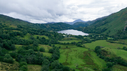 Obraz premium A stunning aerial view of a lush green valley with a serene lake in the distance, surrounded by rolling hills and dense forests under a cloudy sky.