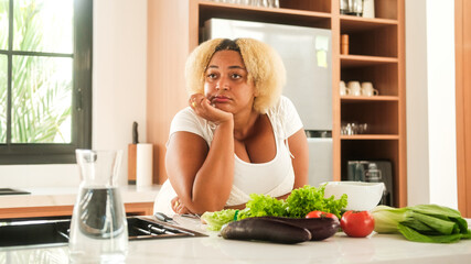 Portrait of a African American curvy woman standing in the kitchen with a tired and sad face and looking at table with vegetables. Depression and fatigue from diets. Self acceptance. Body positivity.