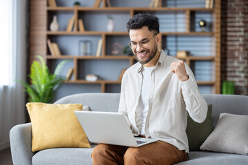 Happy young Indian man sitting on sofa at home, using laptop on lap, looking smiling at screen, showing victory yes with hand