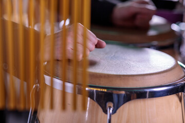 A close-up of a hand playing a conga drum, partially obscured by chimes in the foreground, emphasizing the delicate interplay of rhythm and melody in a percussion setup.