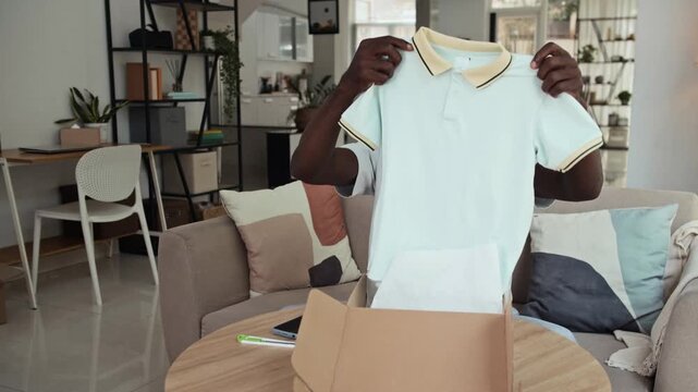 Wide shot of male African American client asking support service of online store about small size of delivered T-shirt using smartphone at home