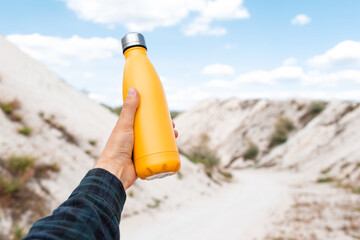 Male hand holding steel thermo water bottle of yellow color on background of sand hills.