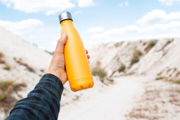 Male hand holding steel thermo water bottle of yellow color on background of sand hills.
