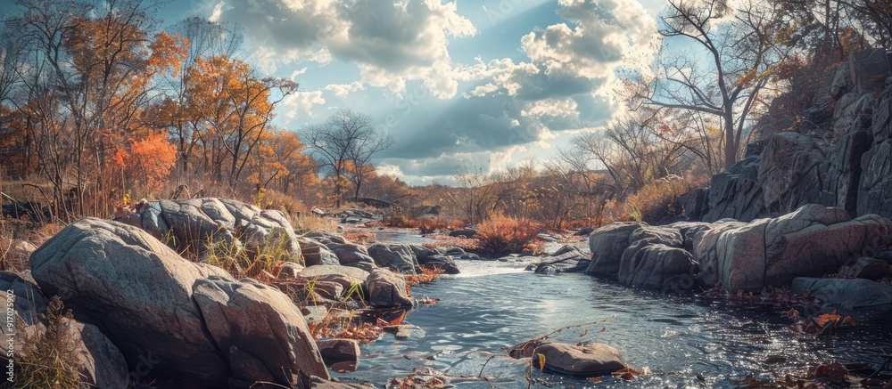 Poster Scenic autumn view with a river cutting through rocks showcasing vibrant fall hues and leafless trees under a sky with fluffy clouds perfect for a copy space image