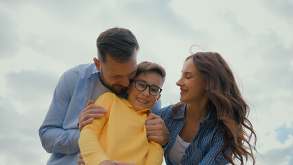 Fototapeta premium Happy family bonding joyful playing Caucasian parents couple mother female woman mom hugging father dad male man guy embracing hug cheerful adopted little boy child kid son outdoors holidays adoption
