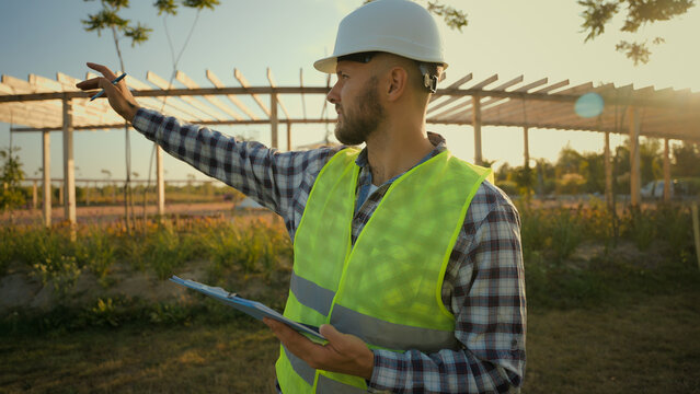 Focused Caucasian man businessman architect male guy engineer hardhat uniform pointing finger showing gesture park checking inspect project writing clipboard looking around construction village meadow