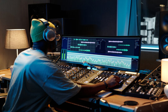 African American music producer working with mixing equipment in well-lit studio filled with various audio devices, wearing headphones and focused on monitors