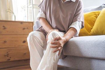 Body arthritis muscles stiff problem, aged woman suffering on leg, pain knee from disease chronic, hand massage rubbing joint injury, mature hurt or sore sitting on sofa at home. Health care people.