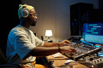 African American man wearing headphones working on music production in modern studio setting. Filled with electronic devices and monitors showcasing editing software © pressmaster