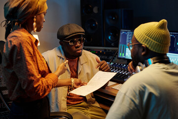 Three musicians actively engaging in music discussion in recording studio, focusing on sheet music and computer screens person wearing headphones sitting at desk