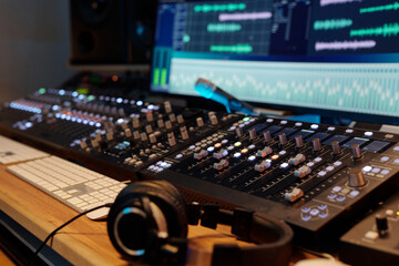 Professional audio mixing board with multiple channels illuminated, showing various sound frequencies on screen in background with headphones resting on desk nearby