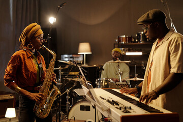 Musicians practicing in intimate studio with saxophone player, keyboardist, and drummer creating...