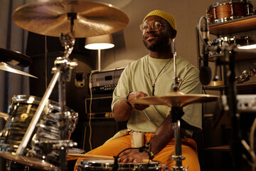 Man with glasses and hat playing drum kit in cozy studio with warm lighting. Surrounded by musical equipment and smiling in concentration