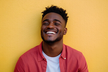 Obraz premium Close-Up Portrait of a Happy Black Man Laughing, Sunlight on His Face, Wearing a Red Shirt Over a White T-Shirt, with Yellow Wall Background