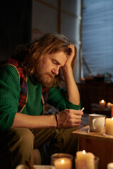 Image depicts long-haired man seated at wooden table, immersed in thoughts, holding a cup surrounded by flickering candles, creating warm ambient atmosphere
