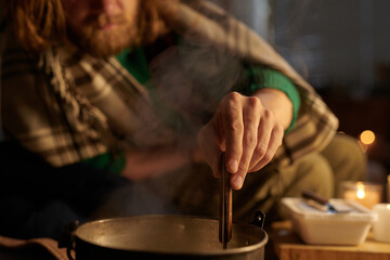 Person stirring soup in pot, wrapped in blanket, with candle in background creating cozy ambiance on cold winter evening