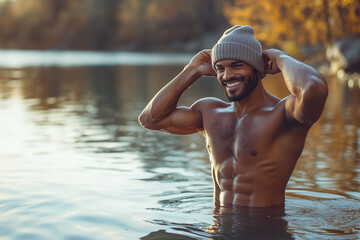 A man is smiling while standing in a calm lake, adjusting his beanie, with the serene early morning or late afternoon light reflecting off the water