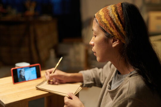 Young woman sketching while watching online art tutorial Using wooden pencil and sketchbook, focused on learning and drawing in cozy indoor setting
