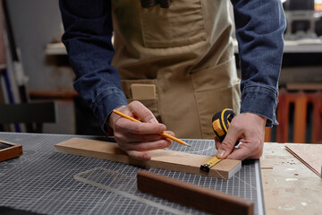 Hands creating detailed measurement on wooden plank using pencil and tool. Person wearing apron and blue shirt in woodworking workshop focusing on perfecting design