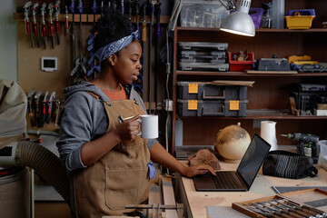 Crafting artisan holding mug in active workshop while working on laptop, surrounded by various...
