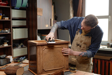 Artisanal woodworker meticulously sanding wooden cabinet in cozy, well-lit workshop, showcasing smooth craftsmanship and attention to detail with assorted tools on workbench